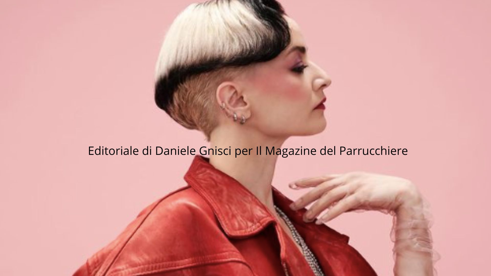 Modella con un taglio di capelli asimmetrico e moderno, in un editoriale di Daniele Gnisci per Il Magazine del Parrucchiere. La modella indossa una giacca rossa e ha capelli corti rasati ai lati con una combinazione di colori biondo platino e nero, su uno sfondo rosa pastello.