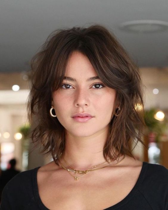 Donna con taglio shag moderno e frangia a tendina, capelli castano scuro con onde morbide, tendenza capelli 2025 per uno stile casual e raffinato.