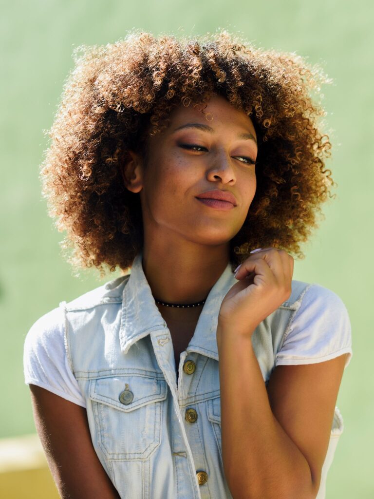 Donna con capelli ricci voluminosi e ben definiti, che guarda con espressione serena, indossando una giacca di jeans senza maniche.