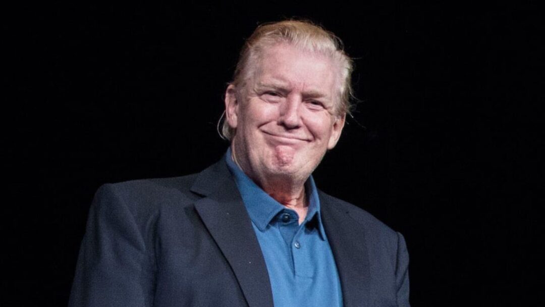 Primo piano di Donald Trump con capelli biondi pettinati all'indietro, che sorride leggermente mentre indossa una giacca scura sopra una polo blu. Sfondo nero.