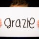 Primo piano di un cartello con la scritta "grazie" tenuto da due mani su sfondo nero, simbolo di gratitudine e apprezzamento.