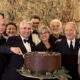 Un gruppo di persone eleganti celebra insieme, posando sorridenti davanti a una torta decorata con cioccolato e motivi natalizi, all'interno di una sala ornata da arazzi.