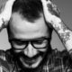 Uomo sorridente con occhiali e tatuaggi floreali sulle braccia.