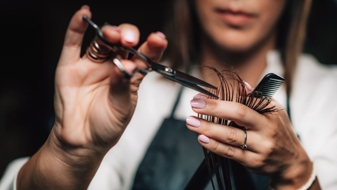 Primo piano delle mani di un parrucchiere mentre taglia con precisione delle ciocche di capelli utilizzando forbici professionali e un pettine nero.