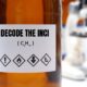 Bottiglia di laboratorio con etichetta "Decode the INCI" e simboli di pericolo chimico, con microscopio sfocato sullo sfondo.
