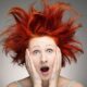 Donna dai capelli rosso acceso in posa sorpresa, con espressione scioccata e mani sul viso. I suoi capelli sono spettinati e voluminosi.