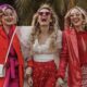 Tre donne sorridenti con outfit rossi e rosa coordinati, acconciature glamour e accessori alla moda.