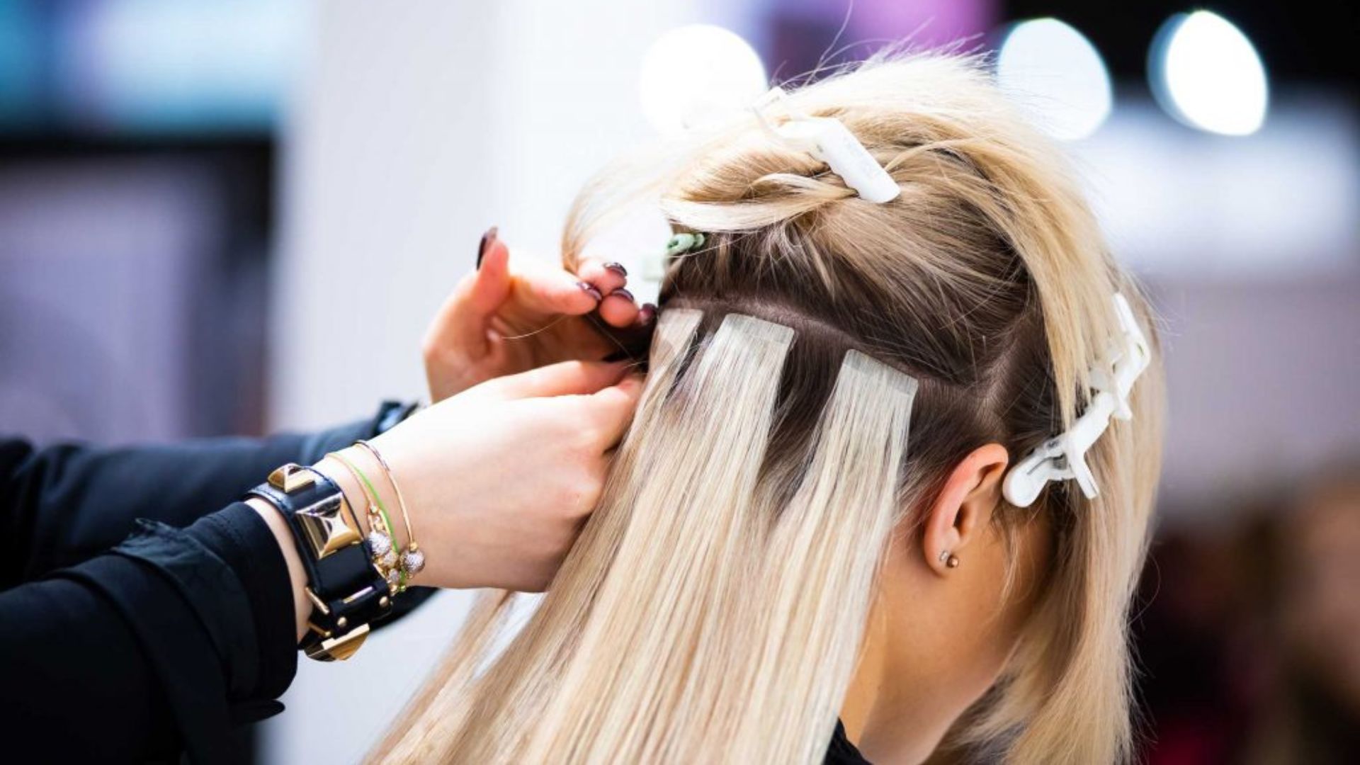 Applicazione di extensions adesive su capelli biondi in un salone professionale. Una hairstylist utilizza clip per sezionare i capelli e fissare le ciocche con la tecnica tape-in, creando un effetto naturale e voluminoso.