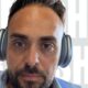 Primo piano di un uomo con cuffie over-ear, sguardo concentrato e camicia chiara. Sfondo con testo sfocato e design moderno.
