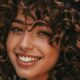 Donna sorridente con capelli ricci naturali tagliati in stile Curly Bob, colore castano caldo con riflessi ramati.