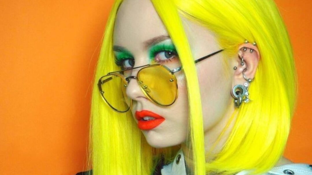 Primo piano di una ragazza con capelli giallo neon, occhiali con lenti gialle, trucco verde acceso e rossetto rosso, su sfondo arancione. Look audace e futuristico in stile Neon Hair Color.