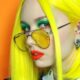 Primo piano di una ragazza con capelli giallo neon, occhiali con lenti gialle, trucco verde acceso e rossetto rosso, su sfondo arancione. Look audace e futuristico in stile Neon Hair Color.