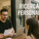 Un parrucchiere intervista una candidata all'interno di un salone, mentre fuori dalla vetrina si vede una fila di persone in attesa; sulla vetrina la scritta "RICERCA PERSONALE".