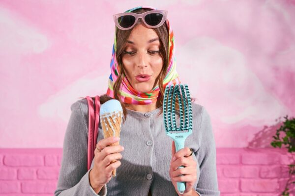 Donna con foulard colorato e occhiali da sole tiene in mano una Fingerbrush Dolce Vita di Olivia Garden e un gelato, su sfondo rosa.