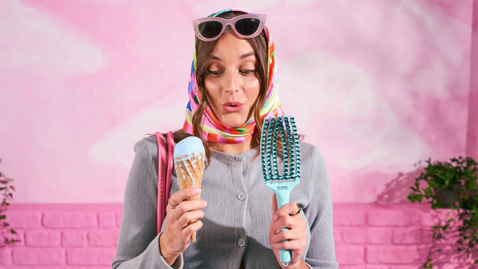 Donna con foulard colorato e occhiali da sole tiene in mano una Fingerbrush Dolce Vita di Olivia Garden e un gelato, su sfondo rosa.