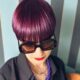 Modella con taglio geometrico corto e colore viola brillante seduta su un divano, con occhiali da sole oversize e sfondo a righe verticali.