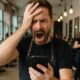 Parrucchiere frustrato con smartphone in mano all'interno di un salone pieno di clienti.