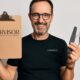 Uomo sorridente con occhiali e barba curata, indossa una maglietta nera con il logo HAIRVISOR, tiene in mano strumenti da manicure e una cartellina, in un contesto professionale.