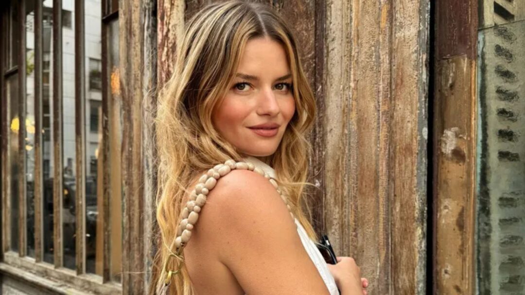 Donna con capelli Jam Blonde ondulati, look naturale e luminoso, posa all’aperto davanti a parete in legno vintage