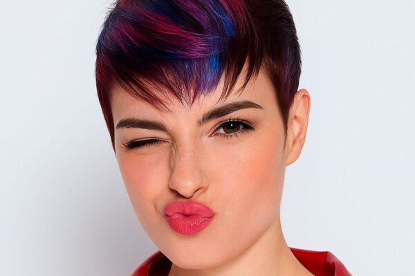 Ragazza con taglio pixie corto e colorazione fucsia-blu, occhi chiusi in un’espressione ironica, giacca rossa lucida