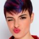 Ragazza con taglio pixie corto e colorazione fucsia-blu, occhi chiusi in un’espressione ironica, giacca rossa lucida
