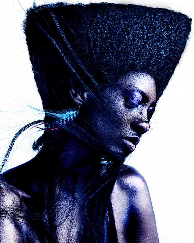 Modella afro con acconciatura piramidale scolpita, piuma turchese tra i capelli e occhi chiusi, pelle effetto granito, su sfondo bianco.