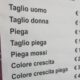 Listino prezzi esposto fuori da un salone di parrucchiere italiano con servizi come taglio, piega e colore, e tariffe aggiornate al 2025.