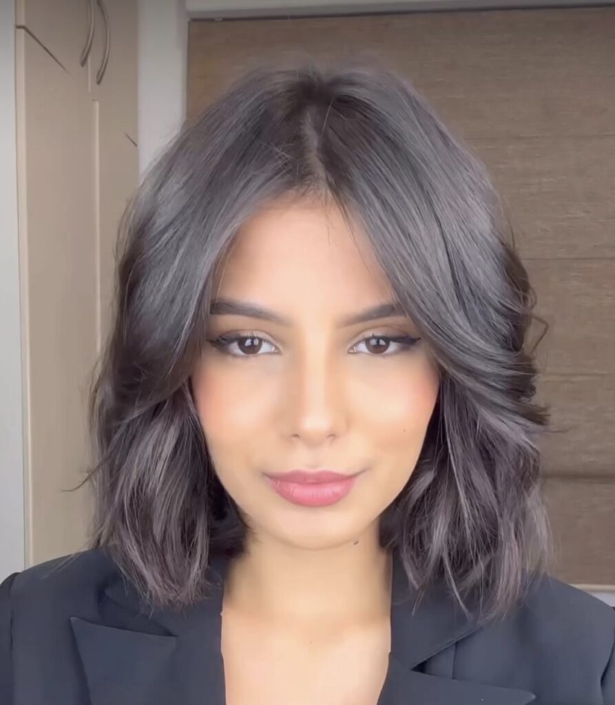 Donna con taglio medio scalato e frangia lunga centrale, capelli castano scuro leggermente ondulati con piega voluminosa e finish lucido.