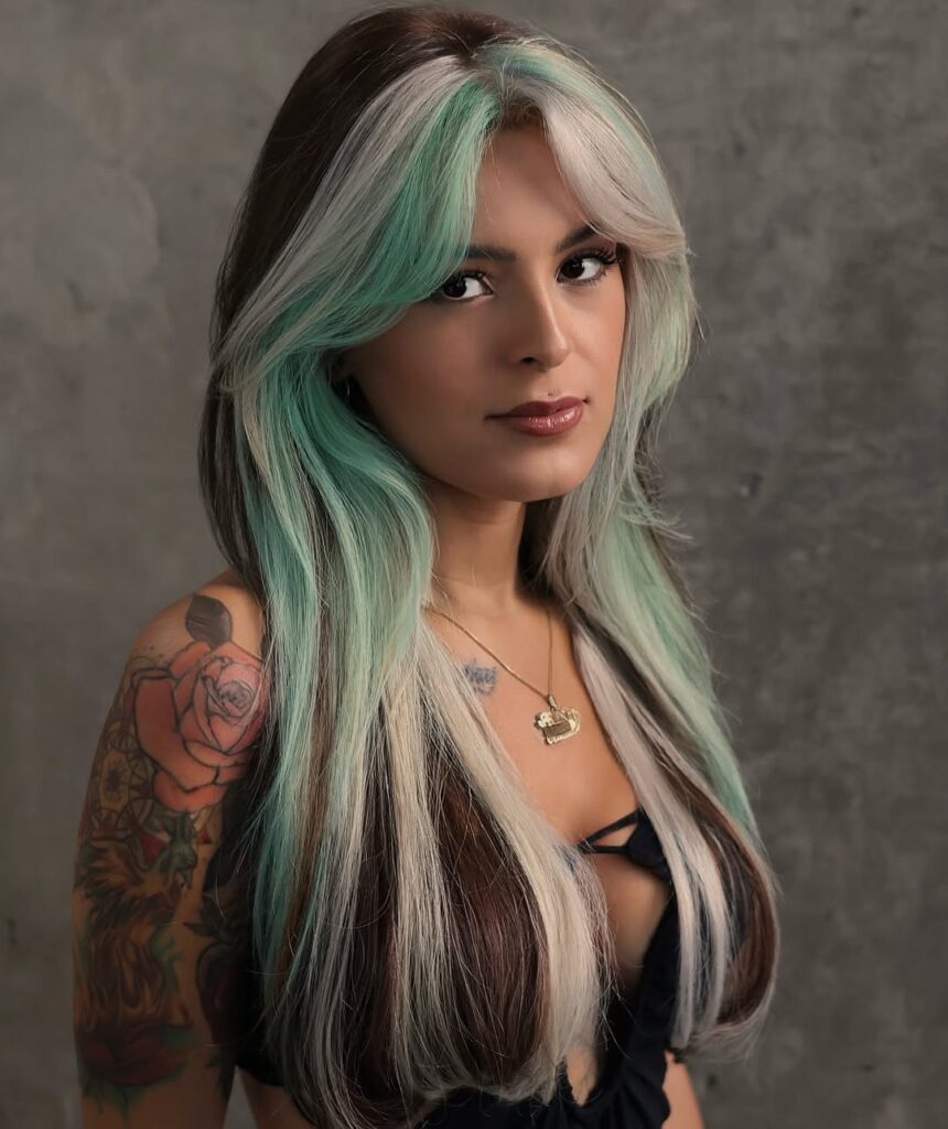 Donna con capelli lunghi color menta, biondo ghiaccio e castano, taglio scalato con frangia a tendina, posa su sfondo grigio cemento.