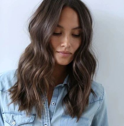 Donna con capelli castani medio-lunghi mossi, riga centrale e styling naturale.