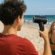 Ragazzo in spiaggia che usa un phon a batteria per asciugarsi i capelli sotto il sole di Ferragosto.