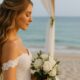 Sposa in spiaggia con capelli a onde morbide half-up, abito in pizzo e bouquet di rose bianche, ritratta davanti al mare.