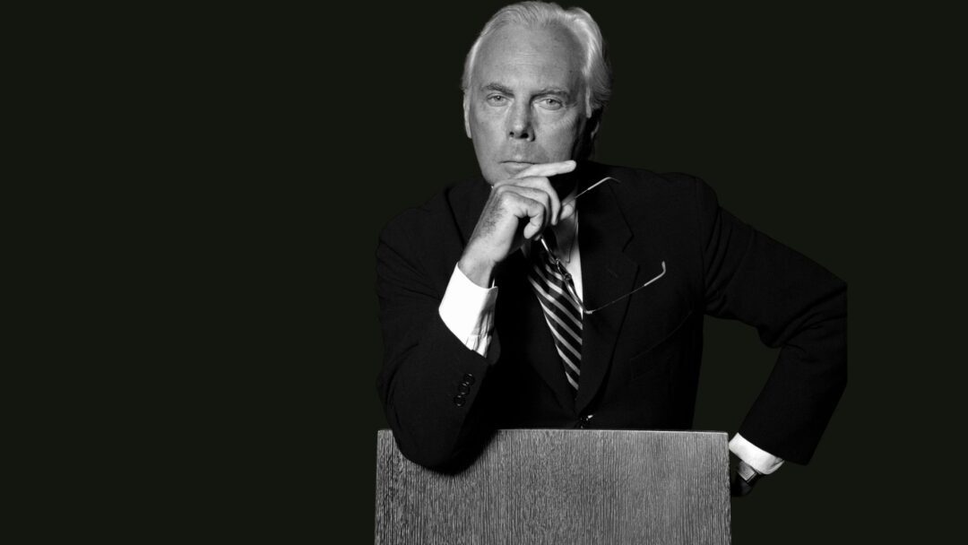 Ritratto in bianco e nero di Giorgio Armani in giacca e cravatta, con posa elegante e sguardo intenso.