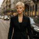 Modella con taglio pixie biondo platino cammina tra le vie eleganti di Milano, indossando un completo nero sartoriale mentre il traffico scorre sullo sfondo.