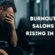 Hairstylist sitting on a barber chair with head down and hand on his face, in a blurred salon environment, representing hairstylist burnout in 2025.