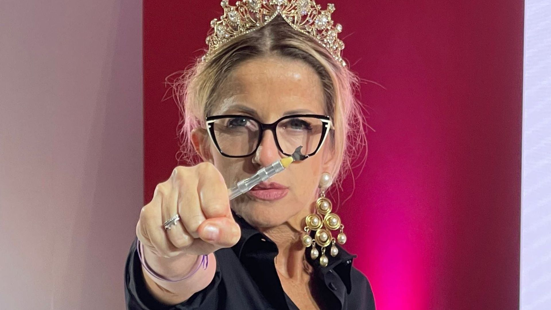 Hairstylist con corona e occhiali punta verso la camera uno strumento tecnico per capelli durante una dimostrazione.