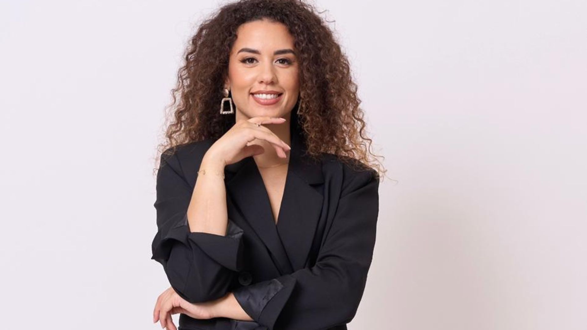 Ritratto professionale di una giovane donna con capelli ricci e castani, che sorride con sicurezza indossando un blazer nero su sfondo bianco.