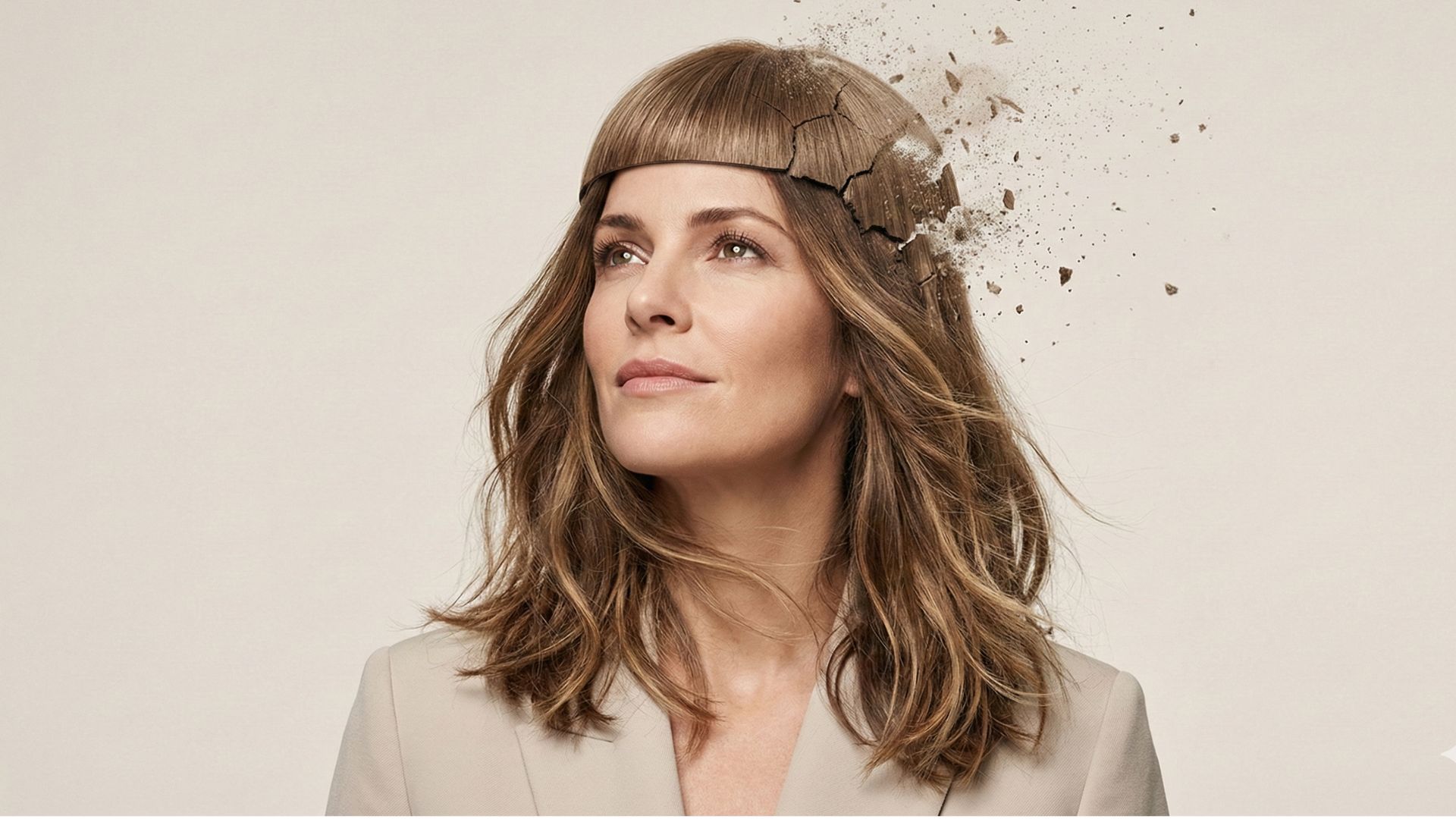 Una donna con espressione fiduciosa guarda verso l'alto mentre una rigida copertura artificiale sulla sommità della sua testa si sta crepando e frantumando in pezzi, rivelando sotto dei capelli naturali e mossi con schiariture moderne. Lo sfondo è neutro e luminoso.