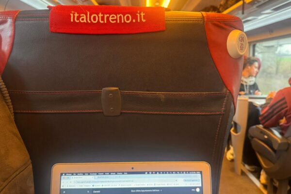 L'interno di una carrozza ferroviaria Italo con un laptop aperto sul tavolino, che mostra una pagina web. Il sedile anteriore ha un poggiatesta rosso con il logo "italotreno.it".