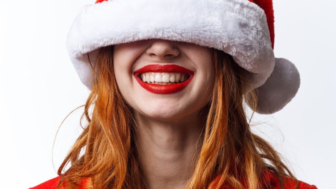 Primo piano di una donna sorridente con capelli rossi che indossa un cappello di Babbo Natale abbassato sugli occhi.