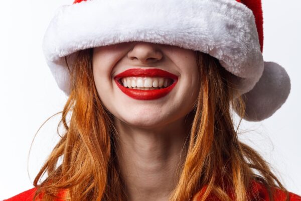 Primo piano di una donna sorridente con capelli rossi che indossa un cappello di Babbo Natale abbassato sugli occhi.