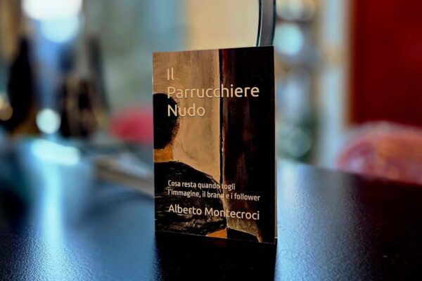 Libro “Il Parrucchiere Nudo” di Alberto Montecroci appoggiato su un tavolo in salone, con sfondo sfocato e luci calde.