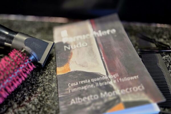 Copertina del libro "Il Parrucchiere Nudo" di Alberto Montecroci appoggiato su un piano di lavoro scuro in un salone, con un asciugacapelli professionale, una spazzola termica fucsia e pettini neri accanto.