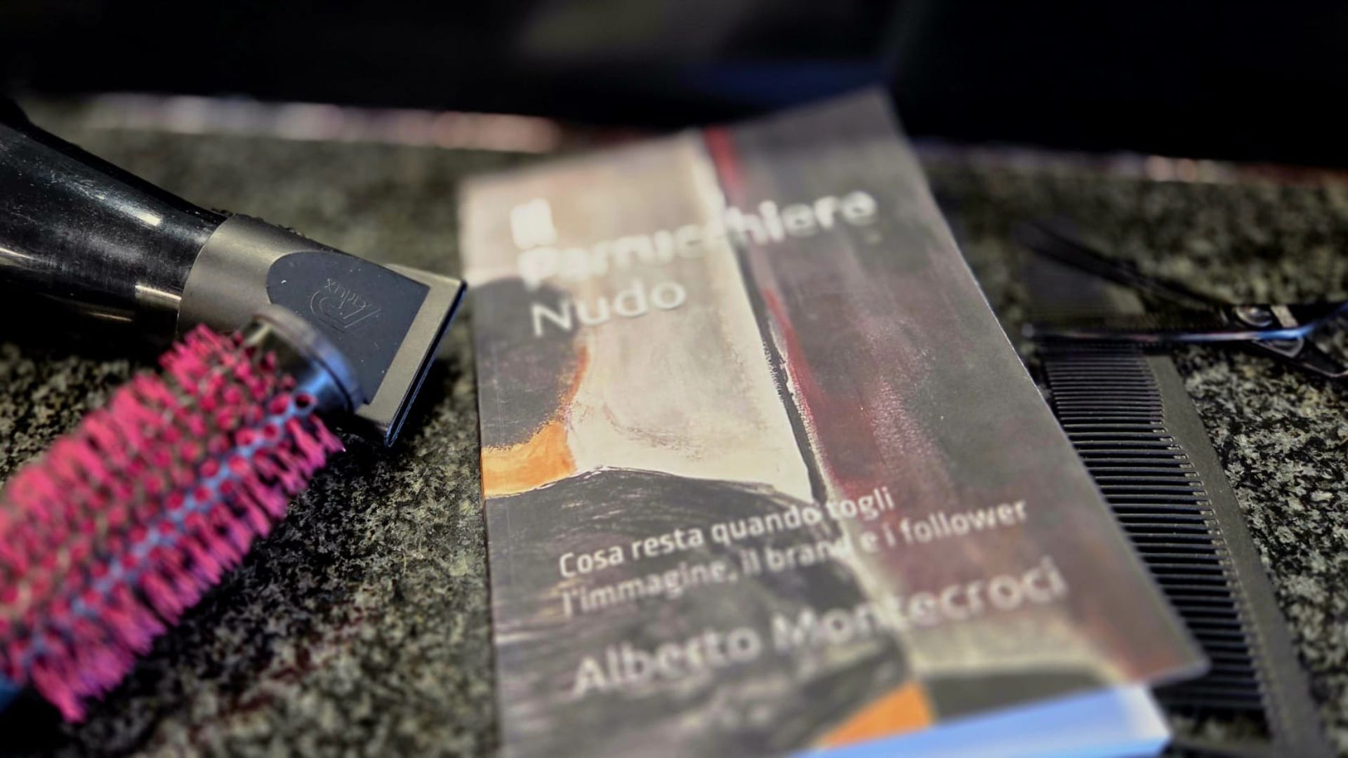 Copertina del libro "Il Parrucchiere Nudo" di Alberto Montecroci appoggiato su un piano di lavoro scuro in un salone, con un asciugacapelli professionale, una spazzola termica fucsia e pettini neri accanto.