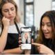 Parrucchiera con espressione rassegnata osserva una cliente che mostra entusiasta sullo smartphone una foto di capelli biondo platino irrealistica e modificata con filtri Instagram.
