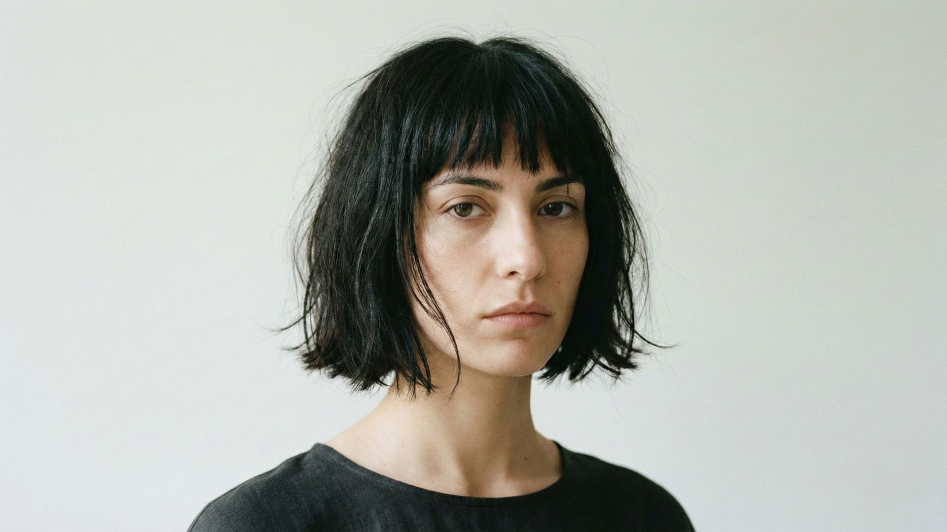 Ritratto frontale di giovane donna con taglio di capelli a caschetto nero stile Japanese Bob spettinato, frangia irregolare e look naturale su sfondo neutro.