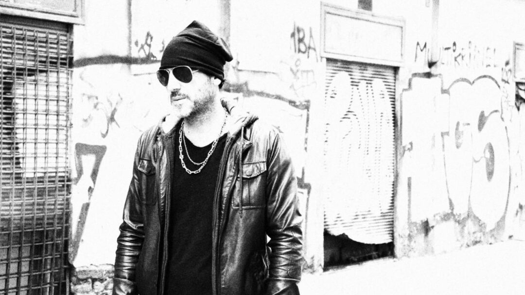 Ritratto in bianco e nero di Antonio Tufano, Hair Architect, in stile urban con giacca di pelle e occhiali da sole, posato di fronte a un muro con graffiti.