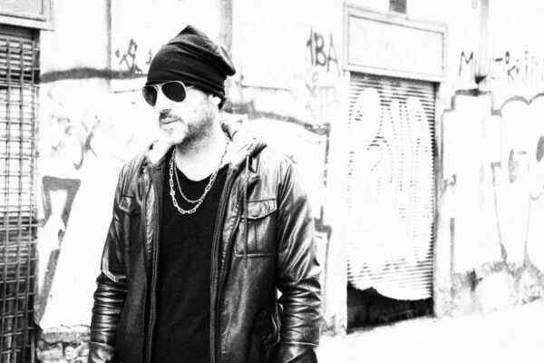 Ritratto in bianco e nero di Antonio Tufano, Hair Architect, in stile urban con giacca di pelle e occhiali da sole, posato di fronte a un muro con graffiti.