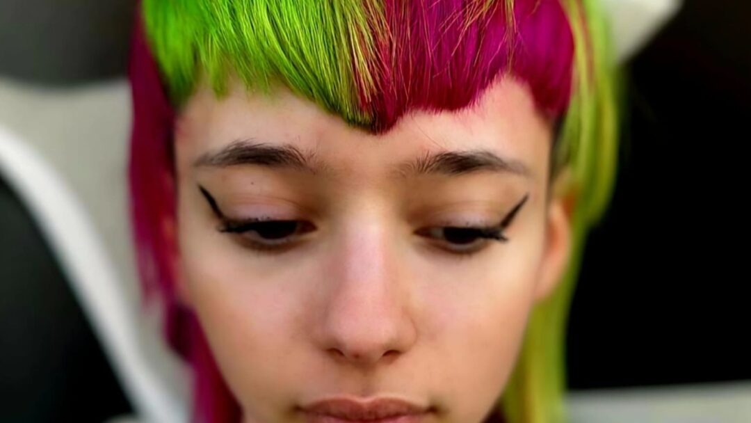 Un primo piano di un viso con un audace trucco occhi grafico nero e capelli corti bicolore verde neon e fucsia brillante. La persona ha gli occhi chiusi e lo sguardo rivolto verso il basso.