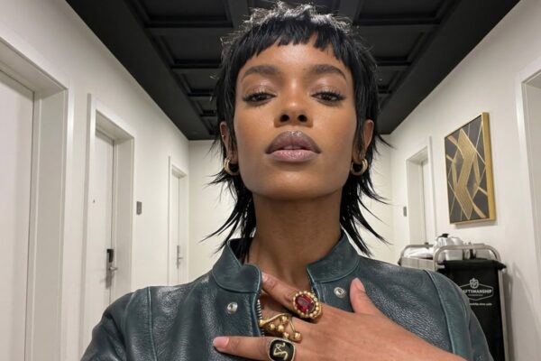 Un selfie iperrealistico a bassa angolazione di una donna afroamericana con un taglio di capelli 'shaggy wolf-cut' (mullet strutturato) nero e testurizzato, che indossa una giacca di pelle verde scuro. Ha la mano sul petto, mostrando un grande anello d'oro oversize con una gemma rossa ovale e altri anelli con simboli neri. Si trova in un corridoio d'hotel minimalista con porte bianche identiche.